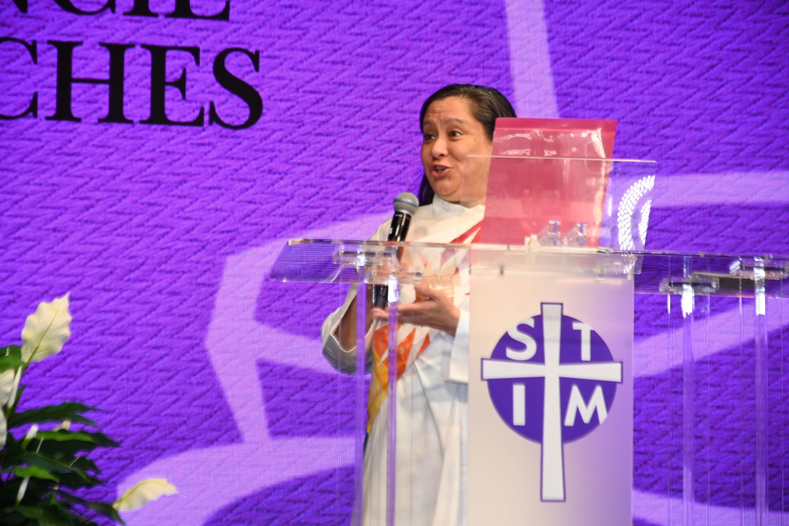 A woman in clergy vestments behind a podium is talking while facing an unseen audience to her right