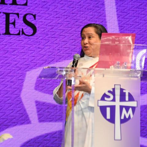 A woman in clergy vestments behind a podium is talking while facing an unseen audience to her right