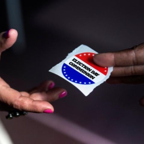 One person giving another person what looks like an "I voted" sticker, but the close-up of the sticker reads "Election Day Communion" instead